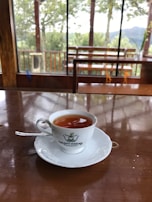 A cup of tea is placed on a white saucer atop a polished wooden table. The cup features a logo for Kelani Valley Plantations. Beyond the glass windows, there is a view of green trees and a wooden railing indicating an outdoor landscape.
