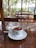 A cup of tea is placed on a white saucer atop a polished wooden table. The cup features a logo for Kelani Valley Plantations. Beyond the glass windows, there is a view of green trees and a wooden railing indicating an outdoor landscape.