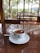 A cup of tea is placed on a white saucer atop a polished wooden table. The cup features a logo for Kelani Valley Plantations. Beyond the glass windows, there is a view of green trees and a wooden railing indicating an outdoor landscape.