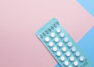 A blister pack containing several small, white pills arranged in rows, placed on a background with pink and blue sections. The packaging seems to be for a form of oral medication, possibly birth control pills.