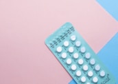 A blister pack containing several small, white pills arranged in rows, placed on a background with pink and blue sections. The packaging seems to be for a form of oral medication, possibly birth control pills.