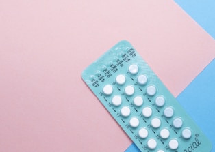 A blister pack containing several small, white pills arranged in rows, placed on a background with pink and blue sections. The packaging seems to be for a form of oral medication, possibly birth control pills.