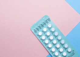 A blister pack containing several small, white pills arranged in rows, placed on a background with pink and blue sections. The packaging seems to be for a form of oral medication, possibly birth control pills.