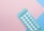 A blister pack containing several small, white pills arranged in rows, placed on a background with pink and blue sections. The packaging seems to be for a form of oral medication, possibly birth control pills.