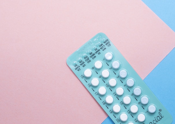 A blister pack containing several small, white pills arranged in rows, placed on a background with pink and blue sections. The packaging seems to be for a form of oral medication, possibly birth control pills.