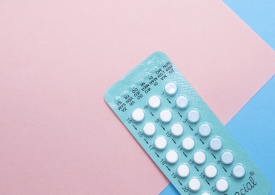 A blister pack containing several small, white pills arranged in rows, placed on a background with pink and blue sections. The packaging seems to be for a form of oral medication, possibly birth control pills.