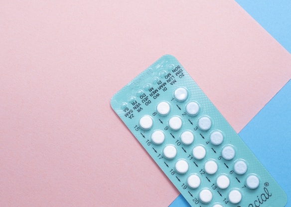 A blister pack containing several small, white pills arranged in rows, placed on a background with pink and blue sections. The packaging seems to be for a form of oral medication, possibly birth control pills.
