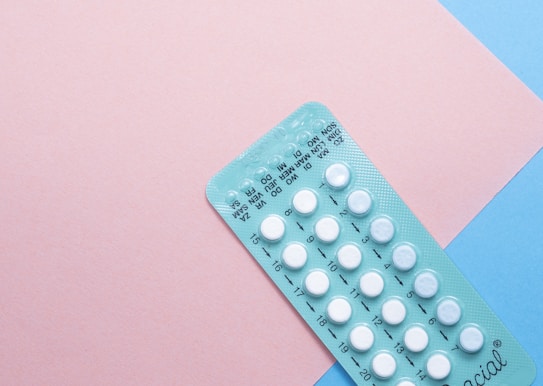A blister pack containing several small, white pills arranged in rows, placed on a background with pink and blue sections. The packaging seems to be for a form of oral medication, possibly birth control pills.