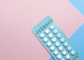 A blister pack containing several small, white pills arranged in rows, placed on a background with pink and blue sections. The packaging seems to be for a form of oral medication, possibly birth control pills.