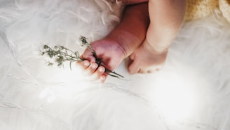 A baby's hand gently holds small white flowers, while the feet rest on a soft, fluffy surface. Warm, glowing lights create a serene atmosphere around the baby, enhancing the delicate and tranquil scene.