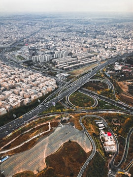 An aerial view of a sprawling urban landscape with a complex network of highways and roads. Residential and commercial buildings are densely packed, with some green spaces interspersed. The roads form intricate patterns with loops and curves, highlighting the city's infrastructure.