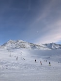 A snowy landscape with a group of people skiing or snowboarding on a ski slope. The background features a mountain range under a clear blue sky with a few clouds.