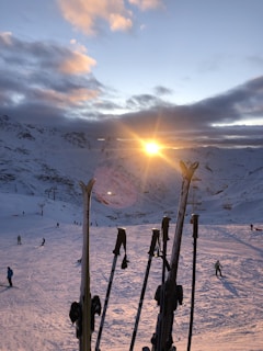 A picturesque snowy mountain landscape with a clear sky partially covered by clouds. The sun is setting or rising between the mountains, casting a warm glow over the snow. Skiers can be seen scattered across the slopes, enjoying the winter sport. Several pairs of skis and poles are propped up in the foreground, adding to the alpine scene.