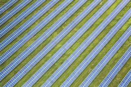 Rows of solar panels are arranged in a diagonal pattern across a green field. The panels are uniformly spaced and cover a large area, emphasizing renewable energy and sustainability.
