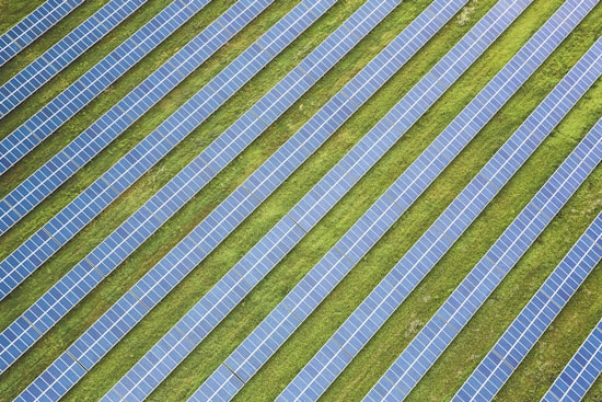 Rows of solar panels are arranged in a diagonal pattern across a green field. The panels are uniformly spaced and cover a large area, emphasizing renewable energy and sustainability.