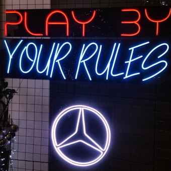 Neon signs on a dark grid background display the phrases 'PLAY BY' in red and 'YOUR RULES' in blue. Below the text, a glowing logo with a circular and triangular symbol is illuminated in white.