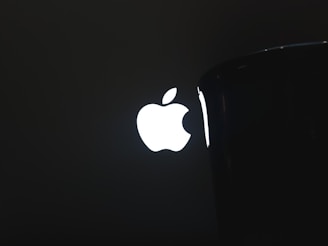 A bright, illuminated logo resembling an apple with a bite taken out of it is prominently displayed against a dark background, partially obscured by an object on the right side.