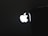 A bright, illuminated logo resembling an apple with a bite taken out of it is prominently displayed against a dark background, partially obscured by an object on the right side.