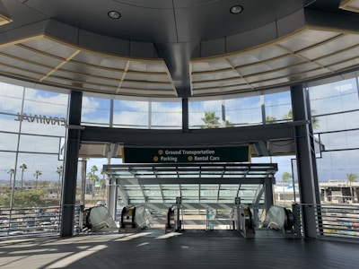 A modern architectural setting with large glass windows providing a view of palm trees and a parking area. The interior has a spacious and open design, featuring escalators leading downwards and signage for ground transportation, parking, and rental cars.