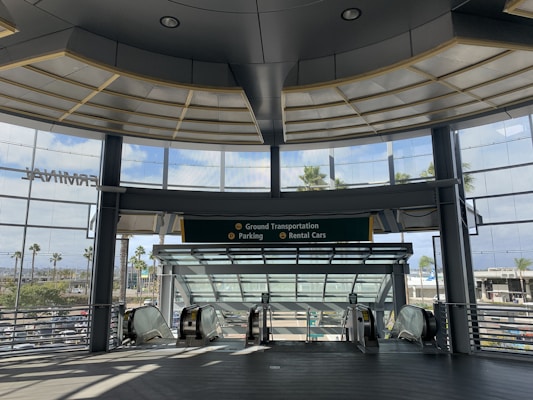 A modern architectural setting with large glass windows providing a view of palm trees and a parking area. The interior has a spacious and open design, featuring escalators leading downwards and signage for ground transportation, parking, and rental cars.