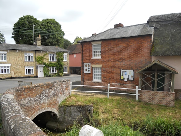 A quaint village scene featuring traditional brick houses with white-framed windows. One of the houses is partially covered in ivy, adding a touch of greenery. An old stone bridge spans a narrow waterway or creek, with grass and foliage surrounding the area. An information board is mounted on the wall of one of the houses, next to a small shelter made of wooden beams with a thatched roof.