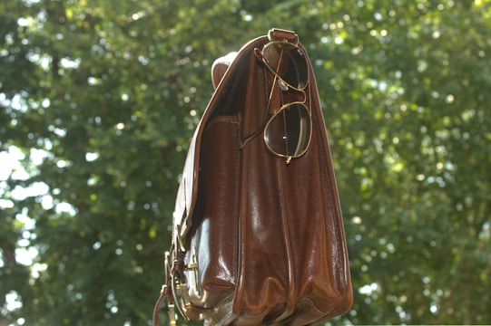 A leather bag hangs in the foreground with a pair of sunglasses dangling from one of its handles. The background consists of lush green foliage, slightly out of focus, creating a natural setting.