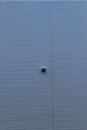 A wall with horizontal corrugated metal panels painted in a muted blue color. A small rectangular light fixture is attached to the center of the wall.