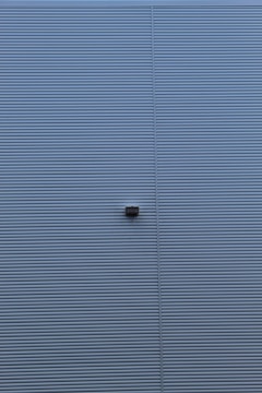 A wall with horizontal corrugated metal panels painted in a muted blue color. A small rectangular light fixture is attached to the center of the wall.