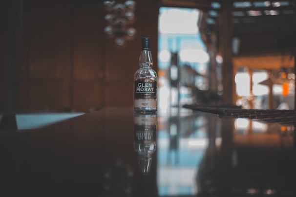 A bottle of Glen Moray whisky sits prominently on a reflective surface, suggesting a bar or lounge setting. The background is softly blurred, creating a sense of depth and focus on the bottle. Warm ambient lighting adds a cozy and inviting atmosphere.