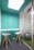 A small, modern office space with turquoise walls and a glass partition. Two white bar stools with wooden legs are positioned at a high, narrow desk. Behind the glass is a large, silver exercise ball on a green, carpeted floor, and a spiral staircase is partially visible.
