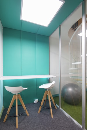 A small, modern office space with turquoise walls and a glass partition. Two white bar stools with wooden legs are positioned at a high, narrow desk. Behind the glass is a large, silver exercise ball on a green, carpeted floor, and a spiral staircase is partially visible.