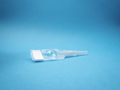 A transparent plastic medical device, likely an eye drop container, is placed against a light blue background. The device is clear and elongated, with a bulbous reservoir at one end and a sealed tip at the other.