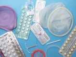 Various contraceptive methods are displayed including birth control pills in blister packs, a diaphragm, a female condom, an intrauterine device (IUD), and an implant. The background is a solid light blue surface.