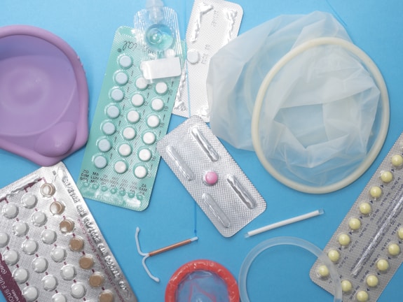 Various contraceptive methods are displayed including birth control pills in blister packs, a diaphragm, a female condom, an intrauterine device (IUD), and an implant. The background is a solid light blue surface.