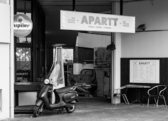 A black and white image captures the entrance of a restaurant named APARTT. A scooter is parked in front of the entrance, alongside a tall outdoor heater. The restaurant signage features bold letters, with additional signs visible for beer and dining options. Outside the restaurant, two metal chairs and a small table are set against the wall.