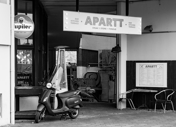 A black and white image captures the entrance of a restaurant named APARTT. A scooter is parked in front of the entrance, alongside a tall outdoor heater. The restaurant signage features bold letters, with additional signs visible for beer and dining options. Outside the restaurant, two metal chairs and a small table are set against the wall.