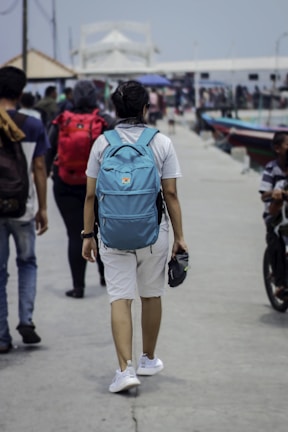 A person wearing a white shirt, white shorts, and a blue backpack walks down a bustling pier. Other people are present, some walking with backpacks, and there are colorful boats on the side. The environment suggests a casual, everyday scene by the waterfront.