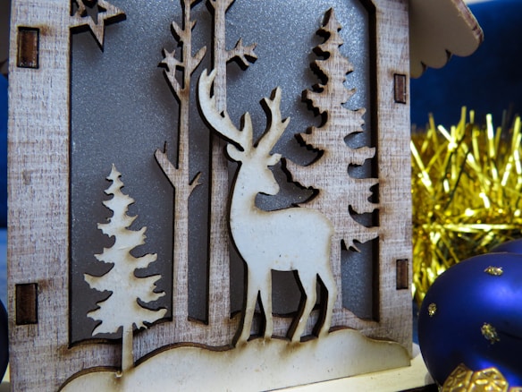 A wooden ornament depicts a reindeer standing among trees in a forest scene. The reindeer is intricately carved alongside simplistic trees, giving a rustic appearance. In the background, a tinsel garland adds a festive, bright touch with gold accents. To the right, a blue Christmas ornament with gold details enhances the holiday atmosphere.