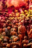 A large crowd of people engaged in a vibrant festival, covered in powders of various colors, predominantly red and yellow. The people are closely packed together, with many wearing similar traditional attire and headwear. Bright colored smoke and dust add to the festive atmosphere.