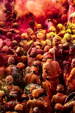 A large crowd of people engaged in a vibrant festival, covered in powders of various colors, predominantly red and yellow. The people are closely packed together, with many wearing similar traditional attire and headwear. Bright colored smoke and dust add to the festive atmosphere.