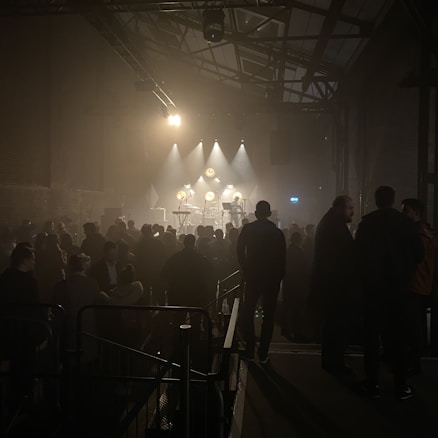 A dimly lit venue with a stage featuring musical instruments and various lighting fixtures. The audience is gathered in front of the stage, casting shadows in the low light setting. The architectural elements of the building, such as metal beams and high ceilings, are visible.