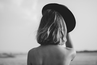 A grayscale photograph featuring the back view of a woman with short hair, holding a wide-brimmed hat. The setting appears to be outdoors with an expanse of open space in the background, creating a sense of solitude or introspection.