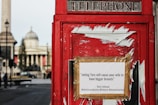 A classic red British telephone box with a satirical poster taped to it. The text on the poster is a political statement attributed to a political figure, alongside hashtags. In the blurry background, historical architecture and a tall column are visible.