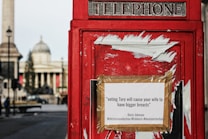 A classic red British telephone box with a satirical poster taped to it. The text on the poster is a political statement attributed to a political figure, alongside hashtags. In the blurry background, historical architecture and a tall column are visible.