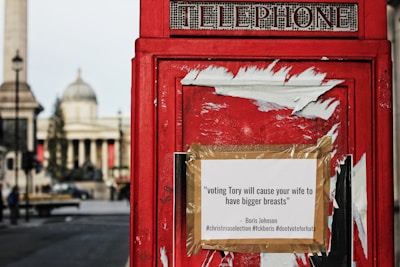 A classic red British telephone box with a satirical poster taped to it. The text on the poster is a political statement attributed to a political figure, alongside hashtags. In the blurry background, historical architecture and a tall column are visible.