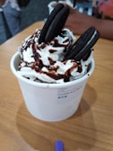 A cup of dessert topped with whipped cream, chocolate syrup, and Oreo cookies sits on a wooden table. The cup's label reads 'Cookies and Creme' with additional text in another language. A hand and another dessert cup are blurred in the background.