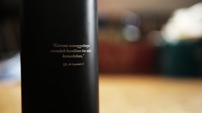 A dark object with a quote inscribed in a lighter color, positioned on a blurry background. The writing appears to be in Indonesian and references a religious text.