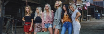 A group of six women standing side by side, smiling and posing for the camera in a rustic, outdoor setting with wooden structures and American flags in the background. They are dressed in a variety of stylish summer outfits, including shorts, skirts, and dresses. The setting gives off a western or country vibe.