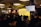 A speaker stands at a podium in a dimly lit room with an audience seated at tables. A large screen displays text about imposter syndrome. The room has industrial-style lighting and brick walls, creating a cozy atmosphere.