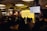 A speaker stands at a podium in a dimly lit room with an audience seated at tables. A large screen displays text about imposter syndrome. The room has industrial-style lighting and brick walls, creating a cozy atmosphere.